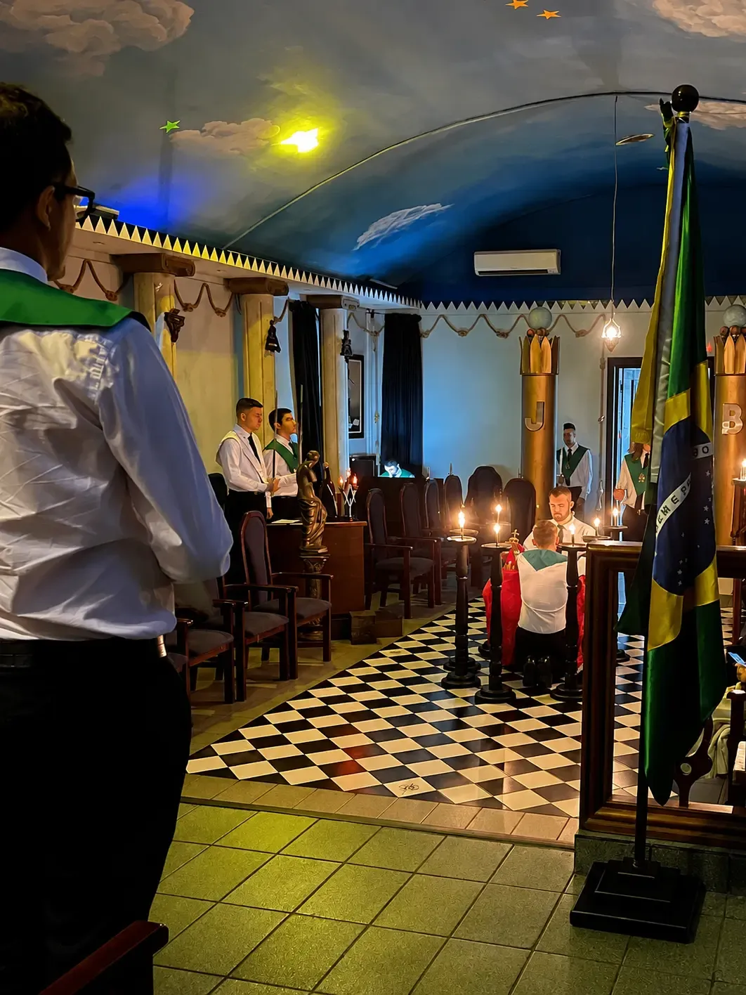 Cavaleiro com faixa verde em um salão à meia-luz, com velas e piso xadrez
