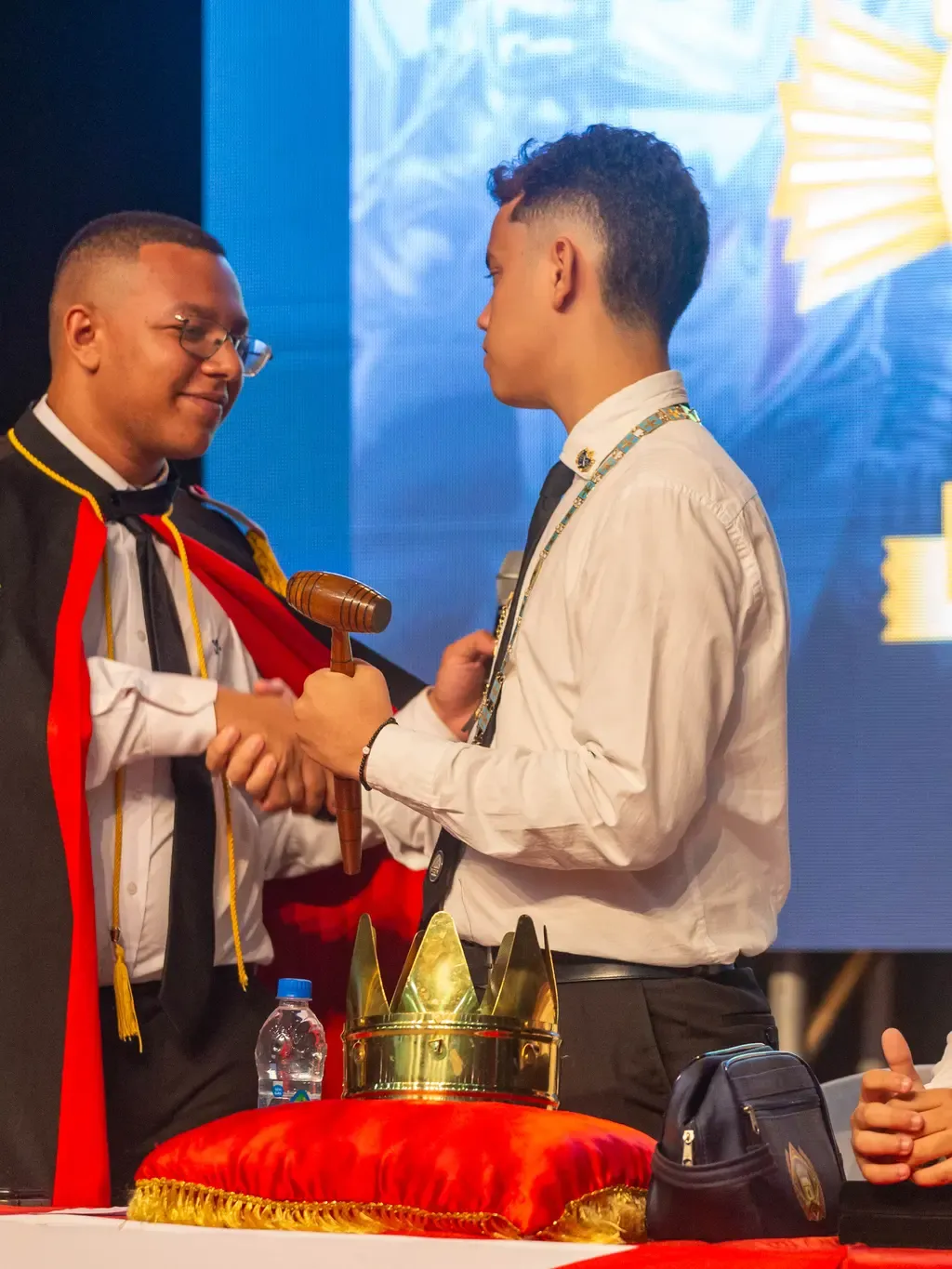 Jovem DeMolay de capa cerimonial cumprimenta um colega no palco durante cerimônia do CEOD