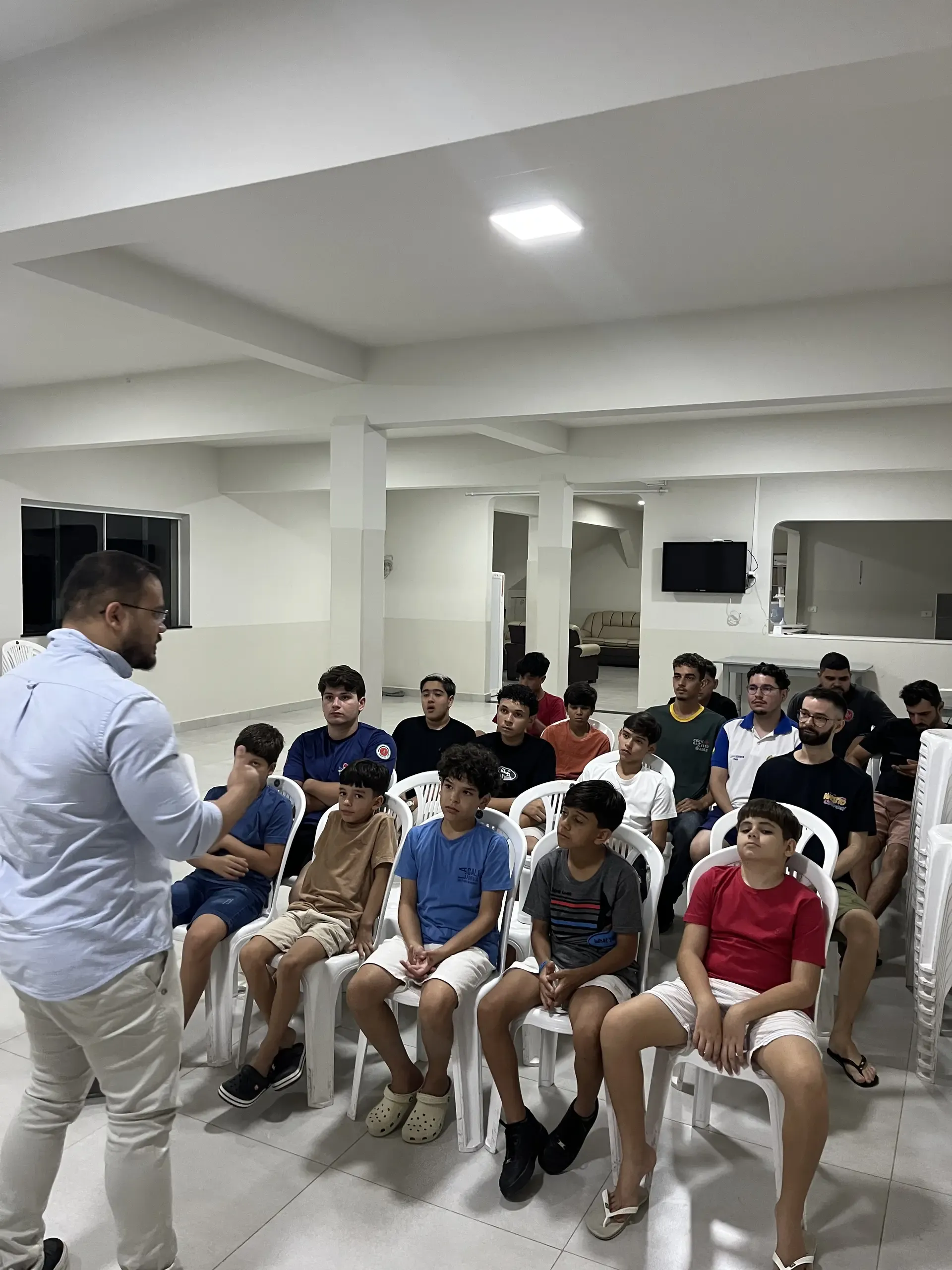 Adulto responsável conversa em pé com os jovens reunidos em uma sala do Capítulo